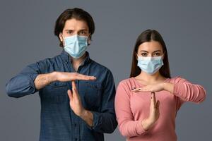 Stop Pandemia. Serious Millennial Couple Wearing Medical Masks Showing Time-Out Gesture While Standing Over Light Studio Background With Copy Space photo