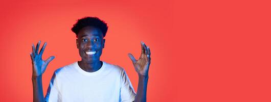 A young black guy is smiling widely and raising their hands in excitement, set against a lively red background. The expression conveys a sense of joy and enthusiasm. photo