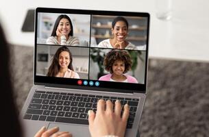 A group of four women smiles at each other during a virtual meeting on a laptop. The setting is bright and cozy, showing a modern workspace. photo