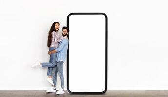 Indian couple stands next to an oversized blank phone display in a well-lit indoor setting, with one partner playfully lifted off the ground, showcasing a fun and dynamic moment. photo