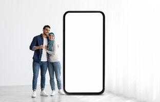 A young Muslim couple stands side by side, beaming with joy while leaning against a large, blank smartphone screen in a bright indoor space filled with natural light. photo