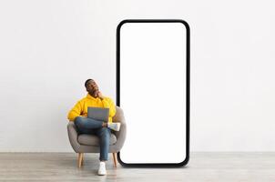 A young black man in a yellow hoodie sits thoughtfully in a chair with a laptop on his lap, beside an enormous blank screen, suggesting a moment of creativity and concentration. photo