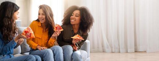 Three multiracial friends laugh and share slices of pizza in a cozy living room. Their joyful expressions highlight a gathering filled with friendship and fun. The space is bright and inviting. photo