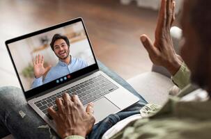 A man is sitting on a couch in his cozy home office, using a laptop for a call. He smiles and waves at the screen, engaged in a friendly conversation with someone. photo