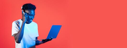 African American young man in a white t-shirt is engaged in a phone conversation while operating a laptop. He smiles, immersed in his work against a bold red backdrop. photo