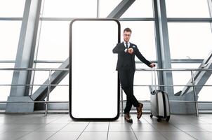 A businessman in a suit stands beside an oversized blank smartphone in a sleek airport terminal. He checks his watch while waiting for his flight, embodying a blend of technology and travel. photo