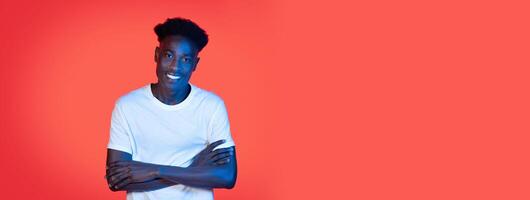 A young black man with a bright smile stands confidently with his arms crossed. The vibrant orange backdrop adds energy, creating a friendly and approachable atmosphere. photo