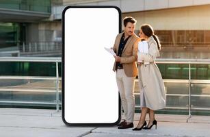 A business couple stands outdoors near a large blank screen, engaging in a conversation while reviewing documents. The sun sets in the background, creating a warm atmosphere. photo