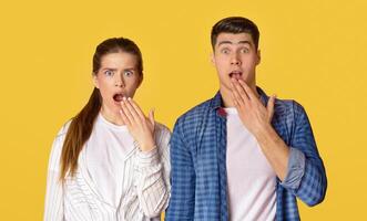 Funny surprised couple see something strange and scared. Excited man and woman look with widely opened mouth at camera and covers it with hand isolated on yellow background, free space, studio shot photo