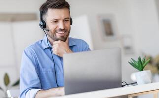 Hotline Support Service. Handsome male call center operator in headset working with laptop in modern office, looking at computer screen, free space photo