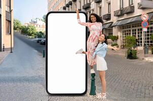 Two friends are having fun in a vibrant city, one standing on a pole and the other next to her, beside a large blank billboard. The surroundings include charming buildings and greenery. photo