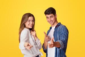 We did it and you can do it. Funny lucky millennial man and woman point fingers at camera and rejoice to success after win at good day, isolated on yellow background, free space, studio shot, cut out photo