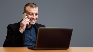 Businessman Working On Laptop Talking On Cellphone Sitting Over Gray Background. Studio Shot, Selective Focus photo