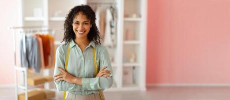 A joyful African American tailor stands confidently in a vibrant clothing store, arms crossed, surrounded by stylish garments on display. The setting is bright, emphasizing creativity and fashion. photo