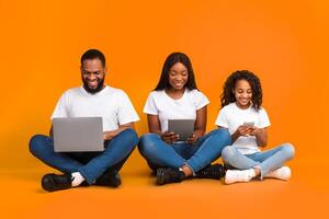 Family Gadgets. Portrait of happy African American parents and their little daughter holding and using different electronic devices while sitting on the floor over yellow studio background photo