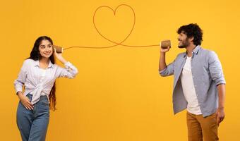 Love message. Loving indian man telling romantic words to girlfriend through tin can phone with heart shaped string, standing over yellow background, creative collage photo