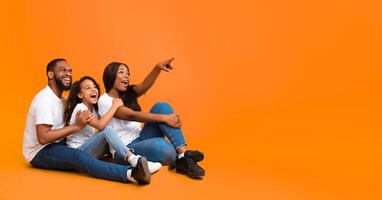 Smiling Black Family Of Three People Sitting On The Floor, Looking Aside, Young Woman Pointing Finger Up At Copy Space On Yellow Background, Demonstrating Free Place For Your Advertisement, Banner photo