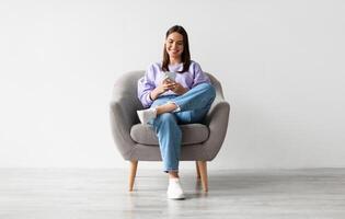 Beautiful young woman sitting in armchair, looking at mobile phone screen, using new cool application against white studio wall, full length. Joyful lady using smartphone for work or entertainment photo