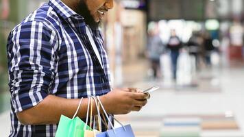 App With Discounts. Smiling Black Man With Shopping Bags Using Smartphone In Mall, Checking Sale Deals, Side View With Free Space photo