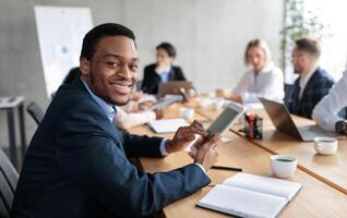 Happy African American Businessman Using Digital Tablet Computer Smiling To Camera Sitting At Corporate Meeting With Colleagues In Modern Office. Business Application Concept. Selective Focus photo
