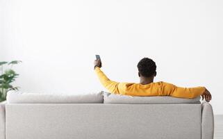 Back view of black man in yellow sitting on couch with remote from TV, pointing towards empty white wall, african american guy turning on air conditioner, home interior, copy space photo