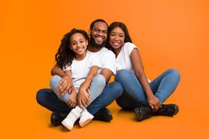 Happy Loving Family. Portrait of African American man, woman and girl sitting on the floor isolated on yellow orange wall. Smiling couple posing with daughter looking at camera. Togetherness, Bonding photo