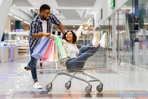 Cheerful black man pulling shopping trolley with his girlfriend inside, having fun while making purchases together in modern department store, fooling and laughing, enjoying seasonal sales, copy space photo