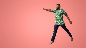 Excited black man in colorful authentic costume running towards copy space for advertisement, blue studio background. Shocked african american guy jumping and pointing at empty space, full length shot photo