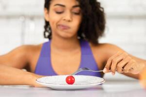 Tired About Diet. Sad african woman looking at one small tomato on a plate, holding fork, not happy at all photo