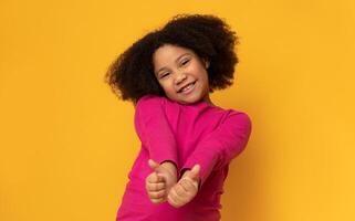 Everything is great. Positive cute little girl gesturing thumbs up and smiling at camera, yellow studio background with empty space photo