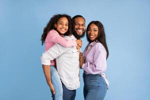Time With Family. Portrait of cheerful African American dad, mum and daughter posing looking at camera standing isolated over blue studio wall. Happy father carrying adolescent girl on back photo