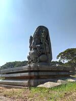 A majestic stone sculpture of Lord Ganesha, intricately carved with a large head and crown, holds an axe and modak. Seated against a clear sky and green trees, it reflects rich Hoysala heritage. photo
