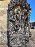 A detailed stone carving of Vishnu, adorns a wall at Chennakeshava Temple. Vishnu stands dynamically, right leg raised, displaying intricate details in garments, jewelry, and physique. photo