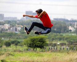 An energetic young man in mid-air performs a martial arts-style jump. Dressed in red and dark pants, he strikes a powerful pose against a grassy field and blurred cityscape, symbolizing energy. photo