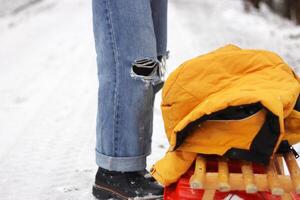 Winter scene with person in ripped jeans and black boots standing near a wooden sled with a yellow jacket on top. Snowy forest path on a cold winter day. Copy space photo