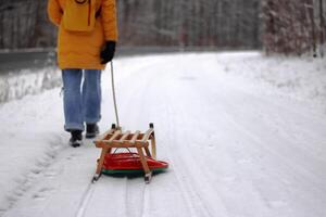 Young woman in yellow jacket pulling a wooden sled on a snowy path by a road. Tenager girl on a winter walk in forest with snow covered trees, Outdoor stroll on a quiet snowy day. photo