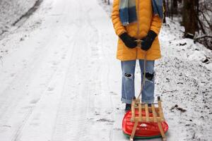 Young woman in yellow jacket pulling a wooden sled on a snowy path by a road. Tenager girl on a winter walk in forest with snow covered trees, Outdoor stroll on a quiet snowy day. photo