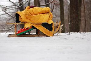 Clolseup of a wooden sled with a warm yellow jacket on a snowy ground in the woods, depicting a winter scene and peaceful retreat into a snow-covered forest. Copy space photo