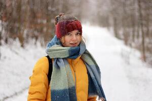 Closeup portrait of a smiling young woman wearing a red knit headband, scarf, and yellow jacket on a cold winter day. Girl dressed warmly surrounded by a blurred snowy forest. photo