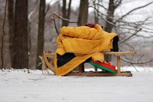 Clolseup of a wooden sled with a warm yellow jacket on a snowy ground in the woods, depicting a winter scene and peaceful retreat into a snow-covered forest. Copy space photo