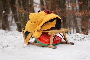 Clolseup of a wooden sled with a warm yellow jacket on a snowy ground in the woods, depicting a winter scene and peaceful retreat into a snow-covered forest. Copy space photo