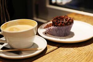 Cozy coffee shop scene with a cappuccino and chocolate muffin on a wooden table by a window in a coffee shop. Selective focus photo