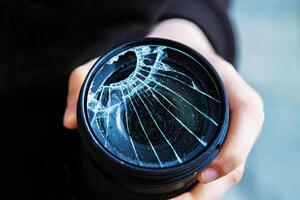 Closeup of a hand holding a broken shattered camera lens with visible cracks, symbolizing damage and fragility of photography equipment, repair or replacement needs, accidents, or fragility. photo