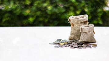 A burlap sack standing and another lying with coins spilling out onto a white surface, set against a blurred background of bushes, representing financial planning and budgeting. photo