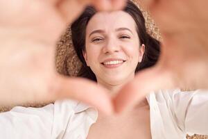 Smiling woman framed by heart-shaped hands, lying down, expressing happiness and positivity in natural light setting. photo