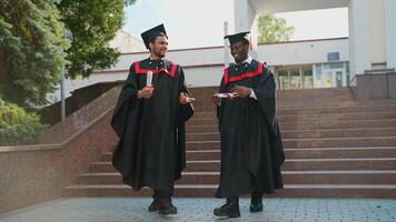 Two multiracial college graduates in graduation mantles walking along university campus and talking video