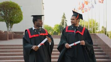 Two multiracial college graduates in graduation mantles walking along university campus and talking video