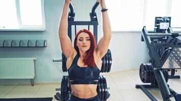 Front view of a beautiful sportive woman who is doing exercises on block simulator. Red-haired athletic female is training her spine muscles on power simulator. Sports workout in the gym. video