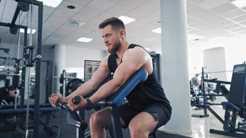 Side view of an athletic man during workout in a gym. Bodybuilder sitting in a modern simulator and trains his muscles of arms. video