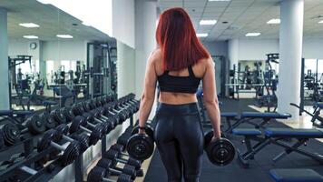 Athletic young woman with perfect body in sportswear lifting dumbbells in the gym. Red-haired female is doing her muscles with dumbbells during training on the background of sports club. video